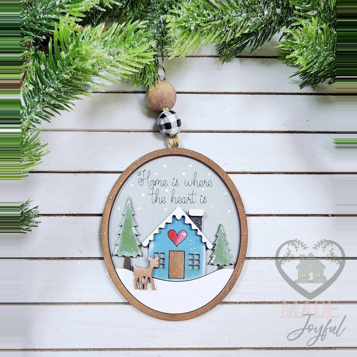 Winter cabin ornament with a blue cabin, heart, deer and pine trees in an oval frame reading Home Is Where the Heart Is, hand-painted in Brentwood, TN.