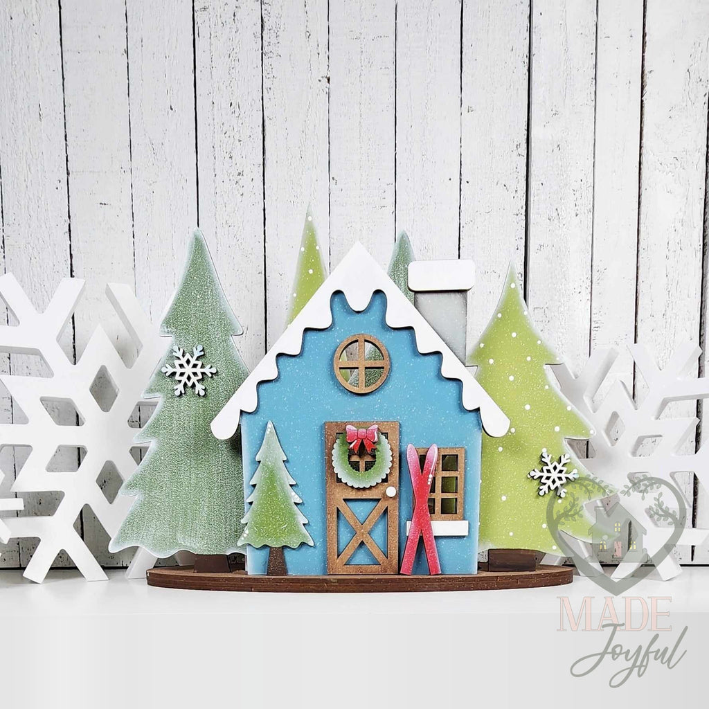 Winter cabin mantel scene with a blue cabin, snowy roof, wreath, and evergreen trees, hand-painted in Brentwood, TN.