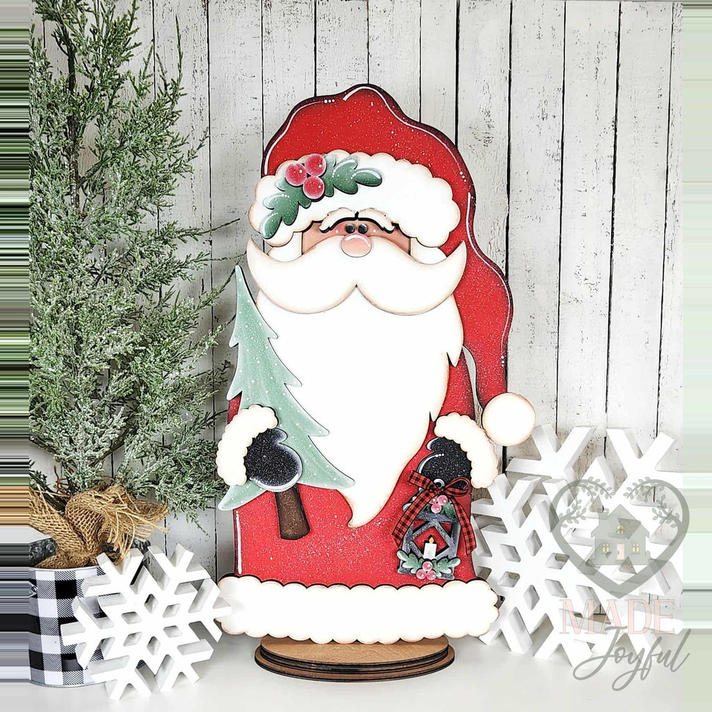Woodland Santa in a red coat holding a tree and lantern, hand-painted in Brentwood, TN.