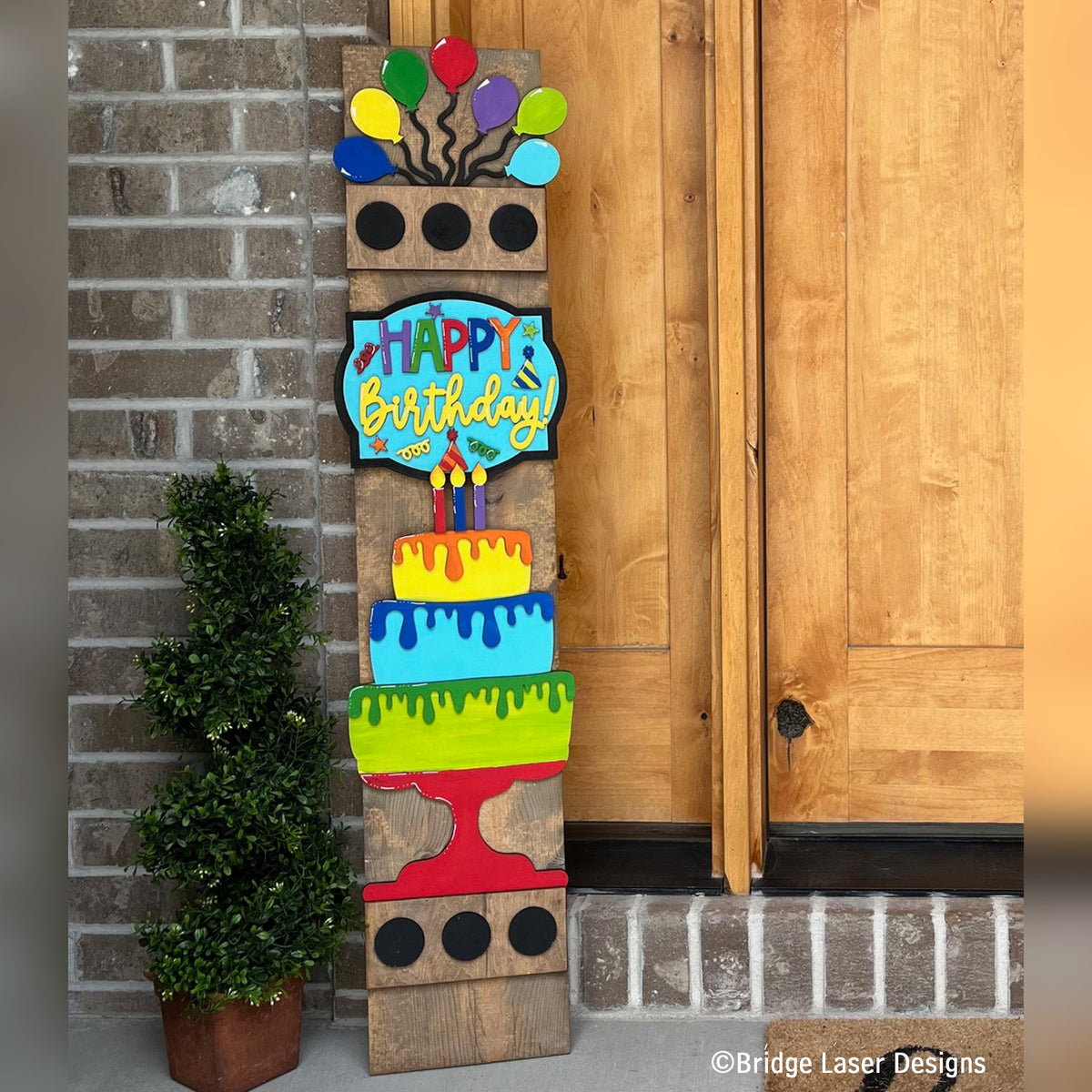 Happy Birthday interchangeable porch leaner with balloons, a Happy Birthday sign, and a layered drip cake on a stained wood base by a front door