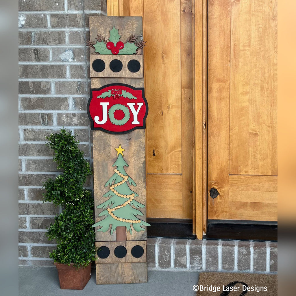 Farmhouse Christmas interchangeable porch leaner with a JOY wreath sign and a green Christmas tree on a stained wood base by a front door