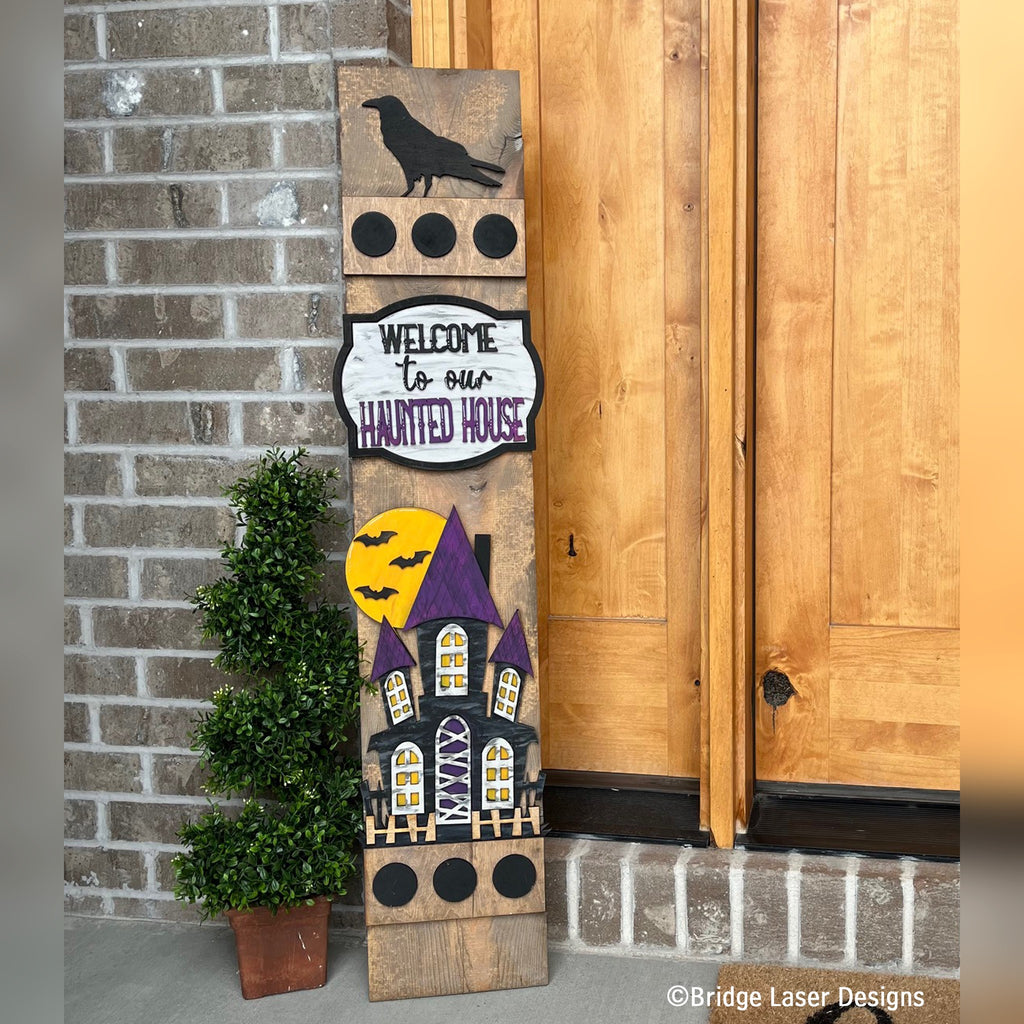 Welcome to Our Haunted House Halloween interchangeable porch leaner with a crow, bats, and a haunted house on a stained wood base by a front door