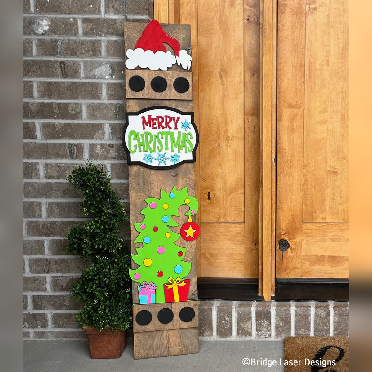 Whimsical Merry Christmas interchangeable porch leaner with a Santa hat, a Merry Christmas sign, and a colorful tree with presents on a stained wood base