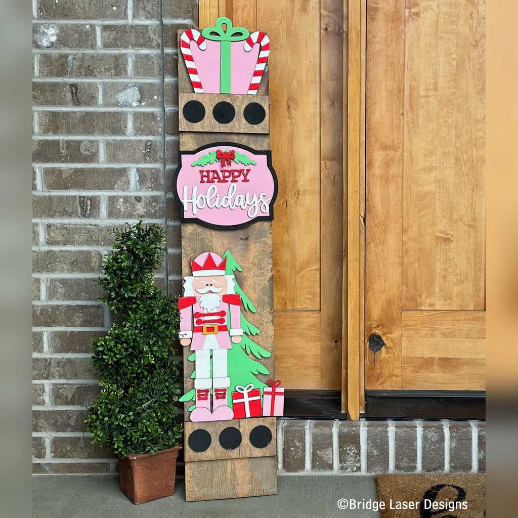 Happy Holidays interchangeable porch leaner with a gift topper, a pink Happy Holidays sign, and a nutcracker with a tree on a stained wood base