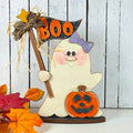 A Halloween-themed wooden ghost decoration holding a small orange jack-o'-lantern and a black sign that says BOO in orange letters. The ghost has a purple bow and pink cheeks, and the decoration is styled with autumn leaves against a white wood-plank background.