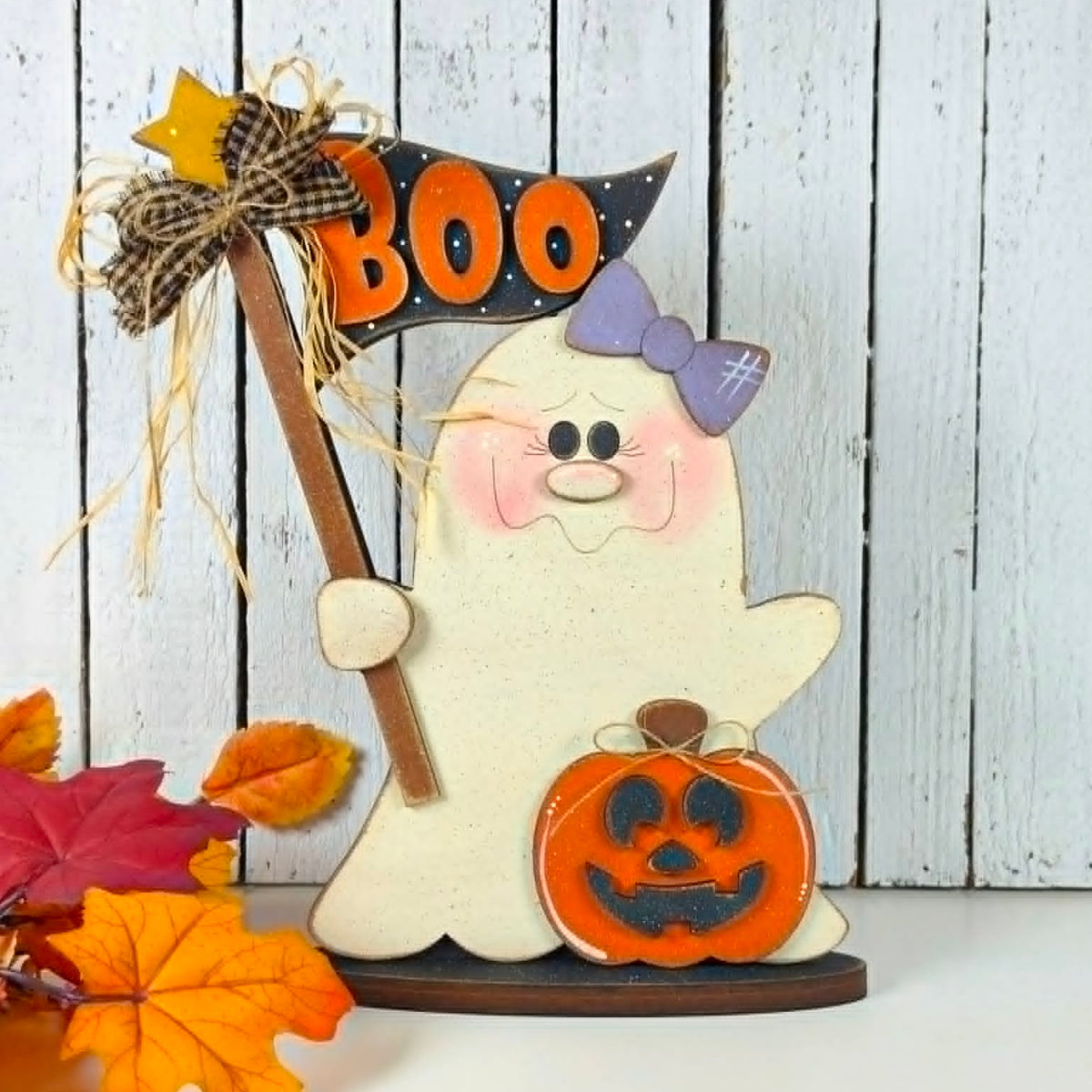 A Halloween-themed wooden ghost decoration holding a small orange jack-o'-lantern and a black sign that says BOO in orange letters. The ghost has a purple bow and pink cheeks, and the decoration is styled with autumn leaves against a white wood-plank background.