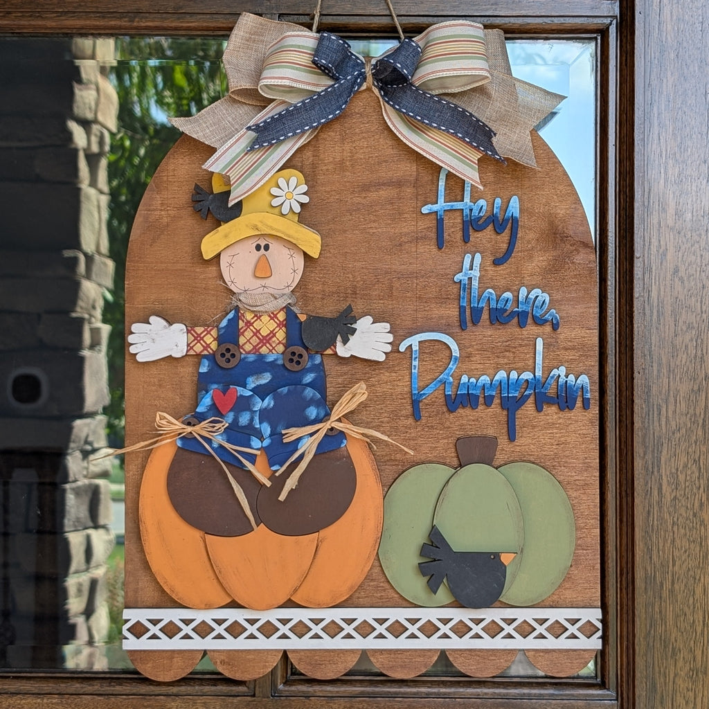 A decorative autumn wooden sign hanging on a glass front door. The sign features a scarecrow sitting on a pumpkin, a green pumpkin, and the phrase 'Hey those Pumpkins' in blue script.