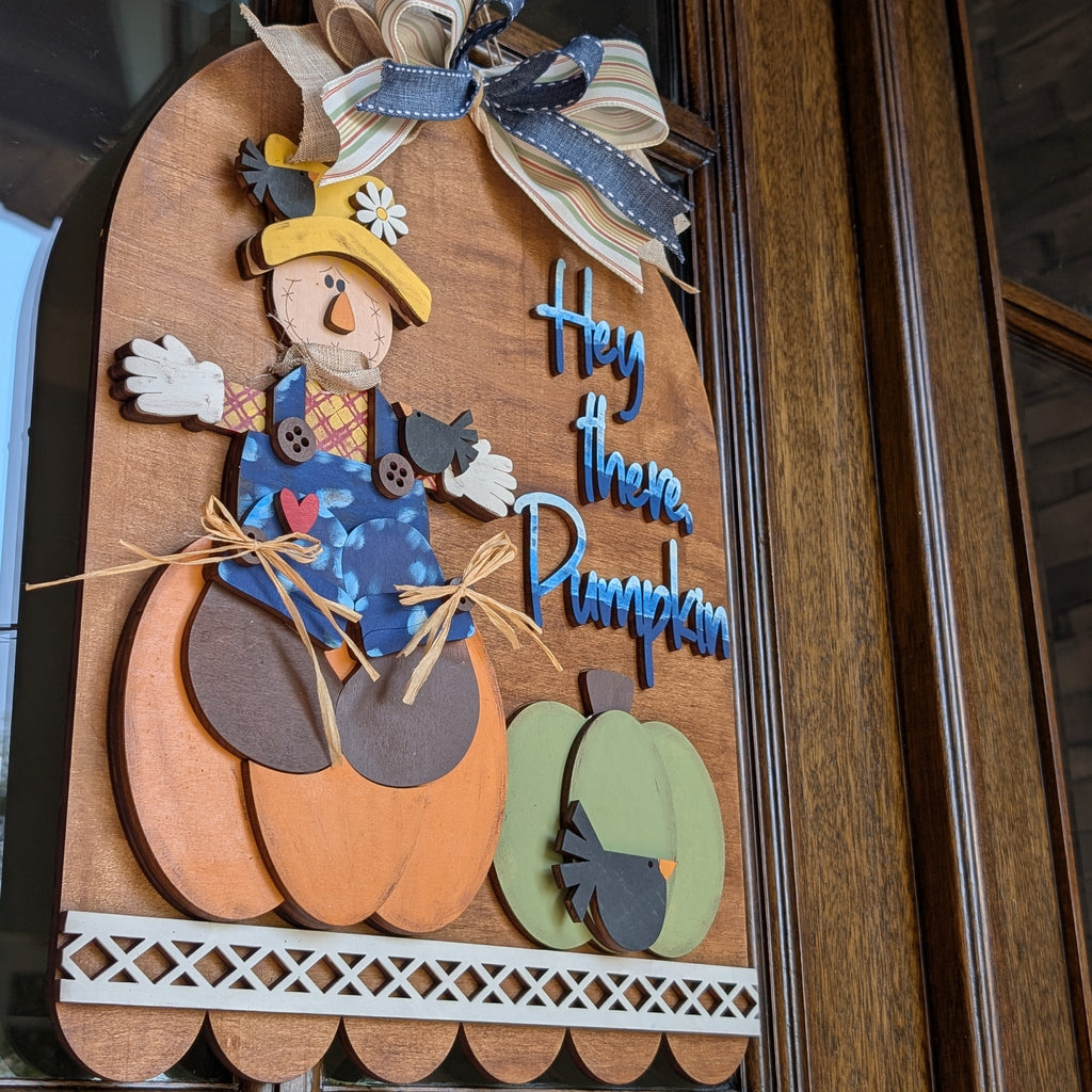 A decorative autumn wooden sign hanging on a dark wood door. The sign features a scarecrow sitting on a large orange pumpkin next to a smaller green pumpkin, with the phrase 'Hey there Pumpkin' in blue lettering.