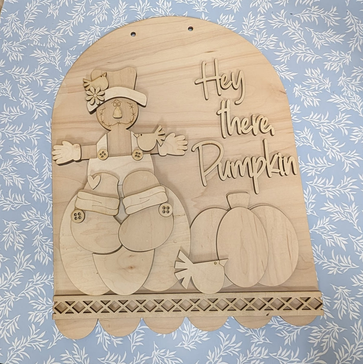 Unpainted wooden autumn wall hanging with a carved scarecrow, pumpkins, bird, and the phrase 'Hey there, Pumpkin' on a blue floral background.