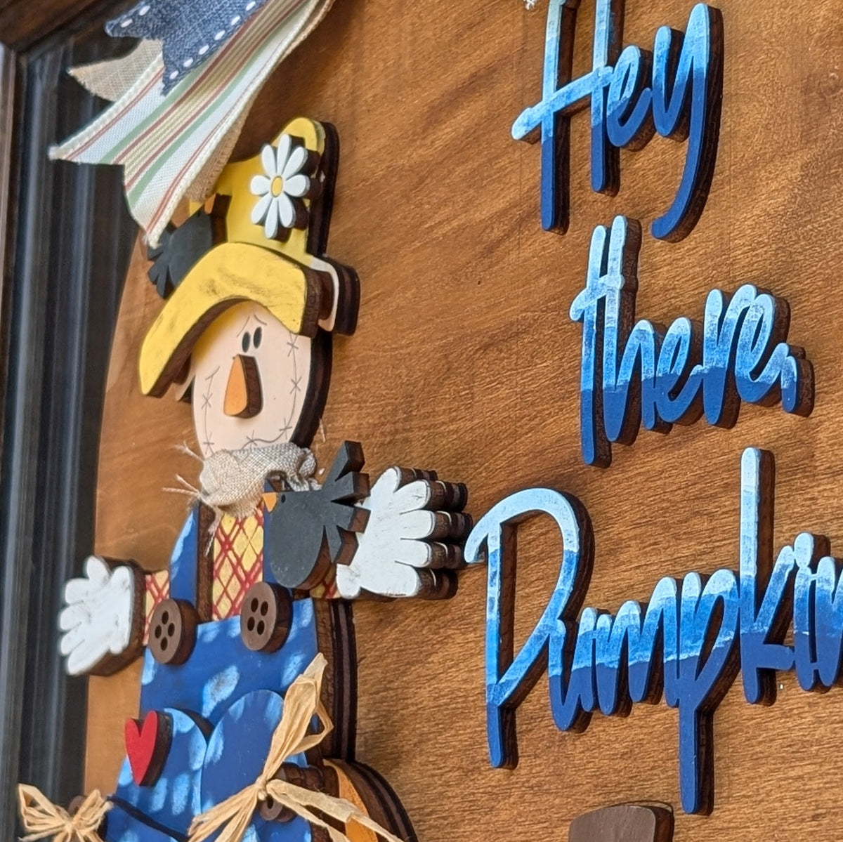 A wooden autumn door sign with the phrase 'Hey there, Pumpkin' in blue script, featuring a scarecrow in a pumpkin, a green pumpkin, and a striped bow at the top, mounted on a dark wood door.