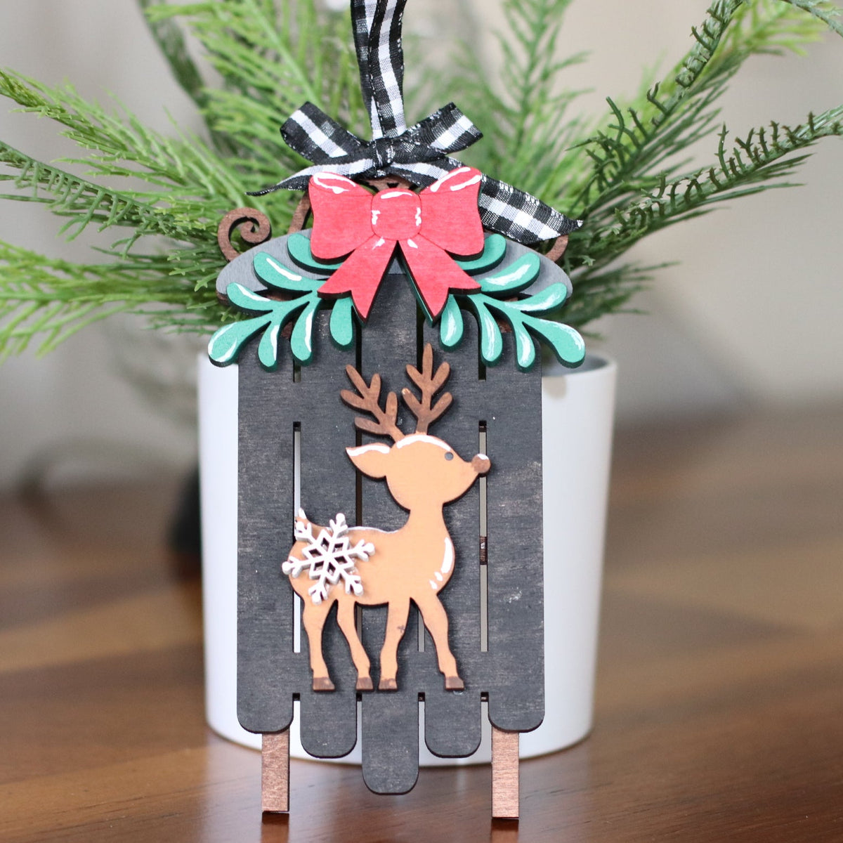 A decorative tabletop ornament featuring a layered wooden deer silhouette over a dark gray picket-style background, topped with a pink bow, teal leaves, and green pine branches in a white pot on a wooden surface.