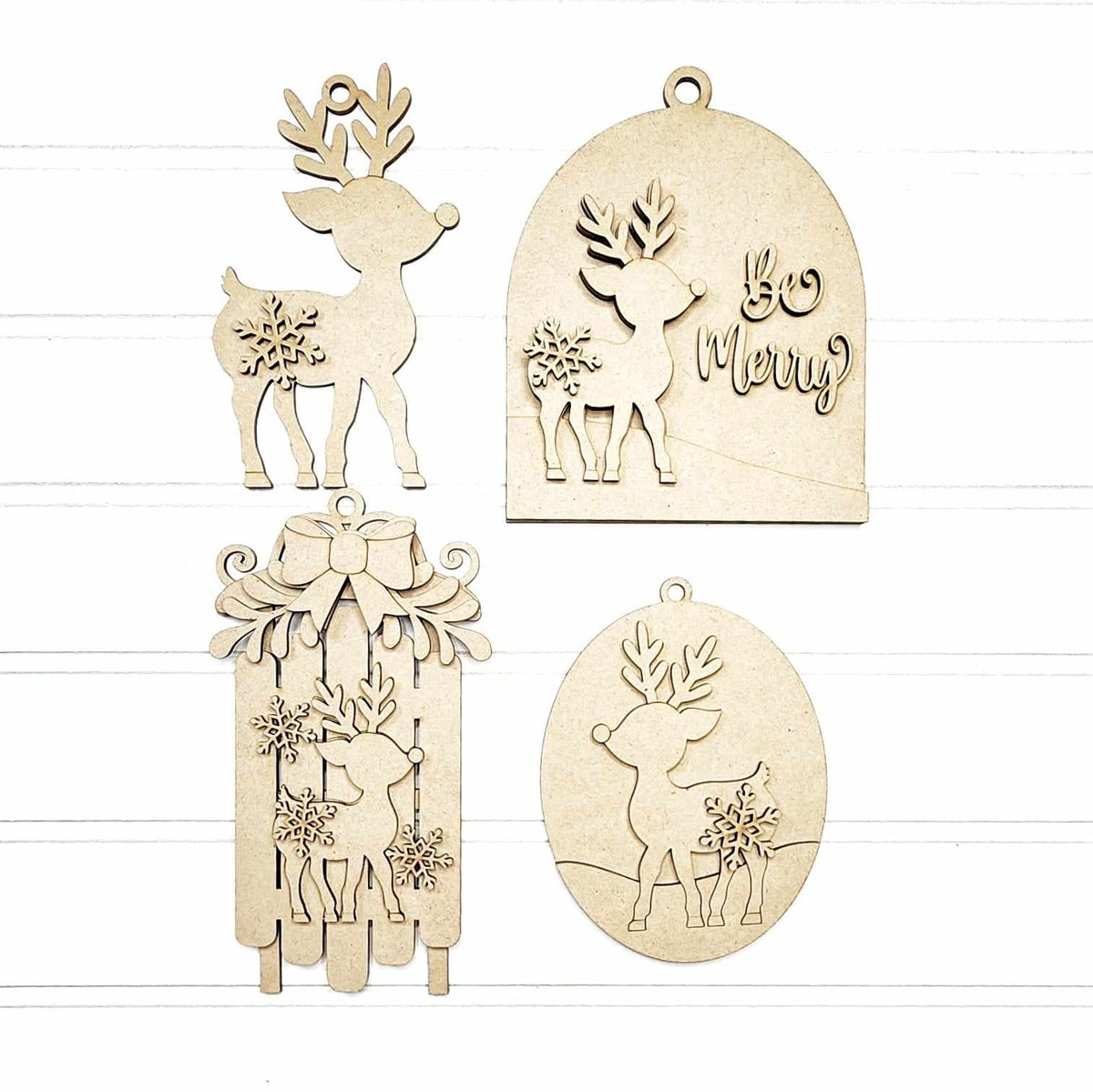 Four natural wood Christmas ornaments with laser-cut reindeer, snowflakes, bows, and 'Be Merry' designs, displayed on a white wood plank background. DIY option