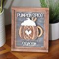 A framed autumn sign with a wooden frame and a dark gray chalkboard-style background. The sign features layered wooden cutouts of a pumpkin mug, sweater pattern with a heart-shaped center, and the words 'PUMPKIN SPICE' at the top and 'SEASON' at the bottom.