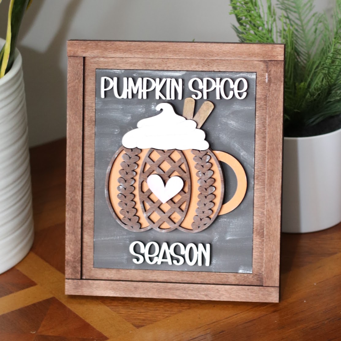 A framed autumn sign with a wooden frame and a dark gray chalkboard-style background. The sign features layered wooden cutouts of a pumpkin mug, sweater pattern with a heart-shaped center, and the words 'PUMPKIN SPICE' at the top and 'SEASON' at the bottom.