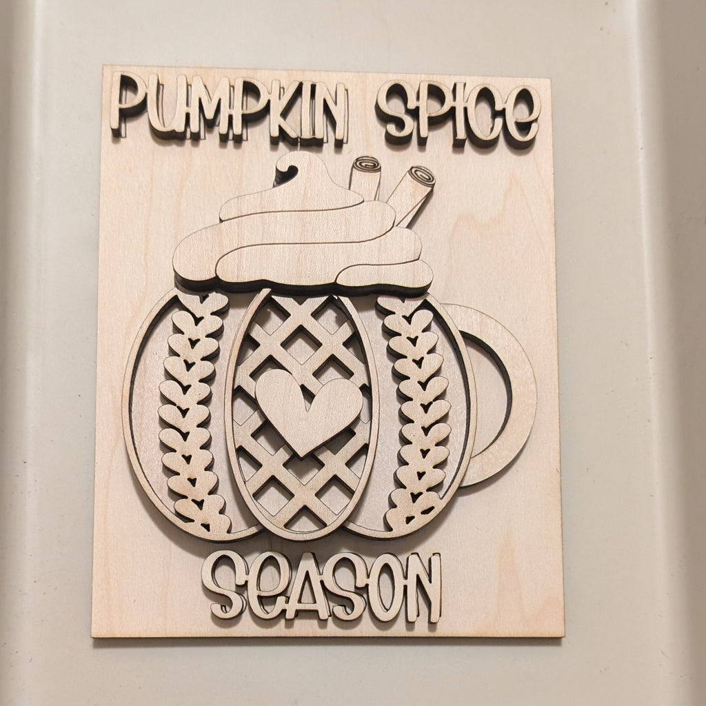  a custom laser-cut wooden sign . The sign features a carved pumpkin mug with sweater pattern a heart in the center, and the text 'PUMPKIN SPICE' at the top and 'SEASON' at the bottom.