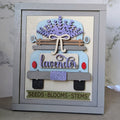 A framed decorative craft piece featuring a stylized lavender truck with layered wood elements, including a bow, blue and lavender truck body, red roundel details, a purple flower crown, and a banner reading 'SEEDS • BLOOMS • STEMS' at the bottom.