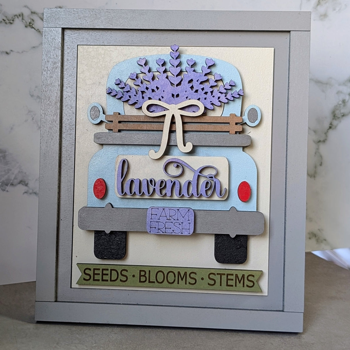 A framed decorative craft piece featuring a stylized lavender truck with layered wood elements, including a bow, blue and lavender truck body, red roundel details, a purple flower crown, and a banner reading 'SEEDS • BLOOMS • STEMS' at the bottom.