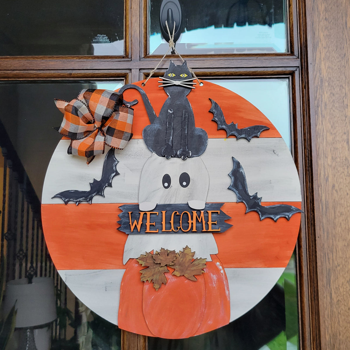 A circular Halloween door wreath hanging on a wooden front door. The wreath features a white and orange striped background with a black cat sitting on a white ghost, black bat cutouts, a plaid orange and brown bow, and a painted orange pumpkin at the bottom with a 'WELCOME' sign across the center.
