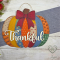 A decorative autumn pumpkin wall hanging with layered patterned panels in orange, blue, green, yellow, and red, topped with a large red bow and the word 'Thankful' in white script. The sign is styled on a white wood surface with a navy gingham ribbon, fall leaves, and greenery.