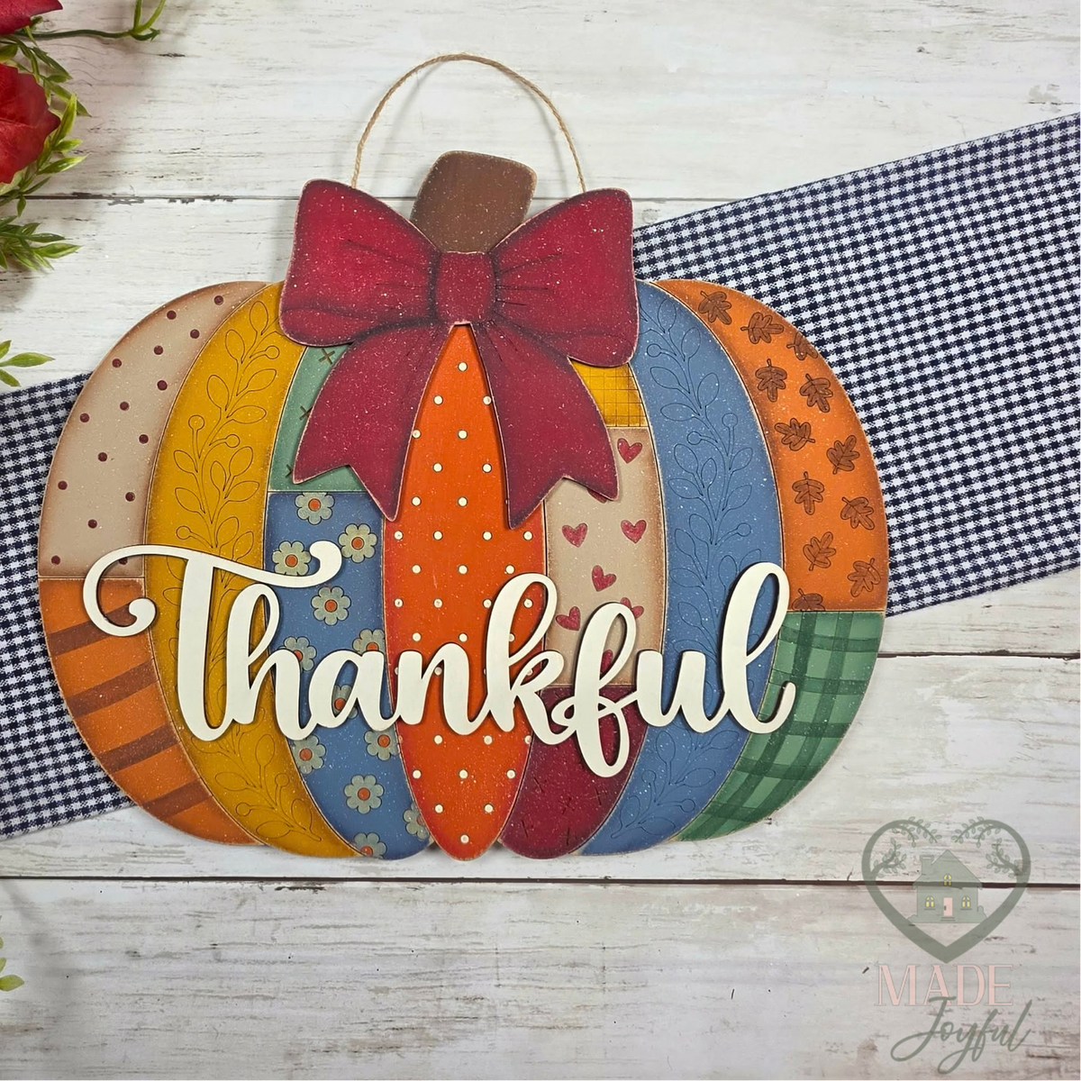 A decorative autumn pumpkin wall hanging with layered patterned panels in orange, blue, green, yellow, and red, topped with a large red bow and the word 'Thankful' in white script. The sign is styled on a white wood surface with a navy gingham ribbon, fall leaves, and greenery.