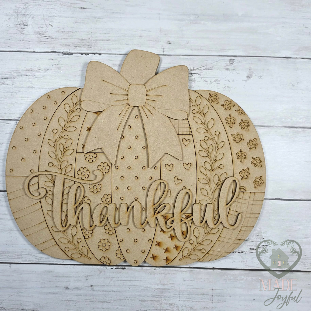 A laser-cut wooden pumpkin sign with a large bow on top, floral vine details, and the word 'Thankful' in script across the front, displayed on a white wood plank background.