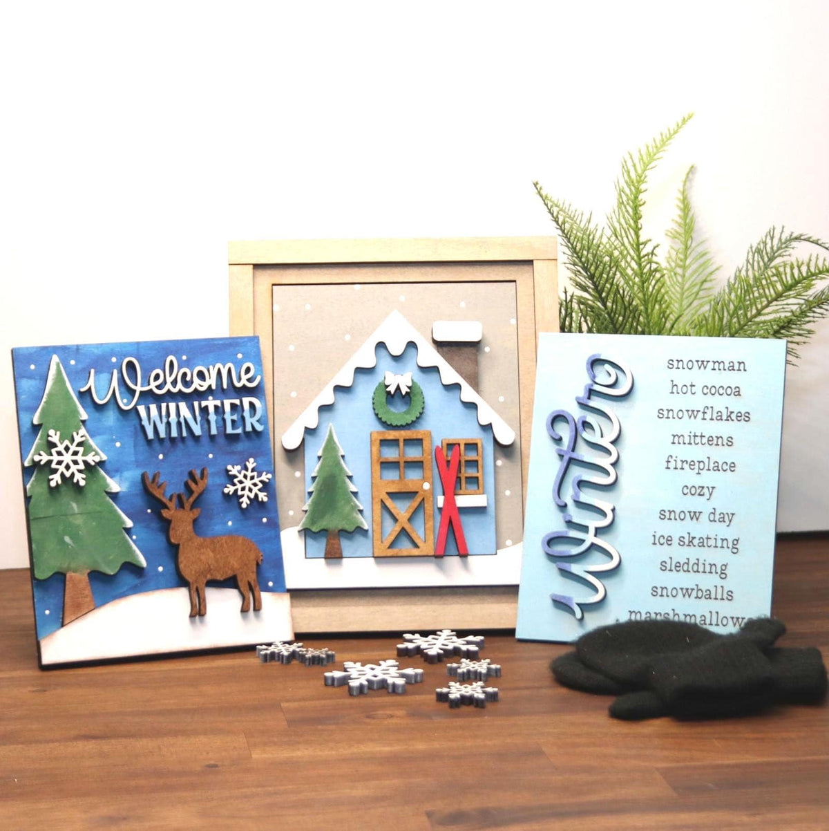 Three handmade winter-themed inserts for 5.5x6" frames arranged on a wooden surface with small snowflake cutouts and a black glove. The left insert says 'Welcome Winter' with a blue background, green tree, white snowflakes, and a brown deer. Perfect for DIY parties in Brentwood  Williamson County, TN areas