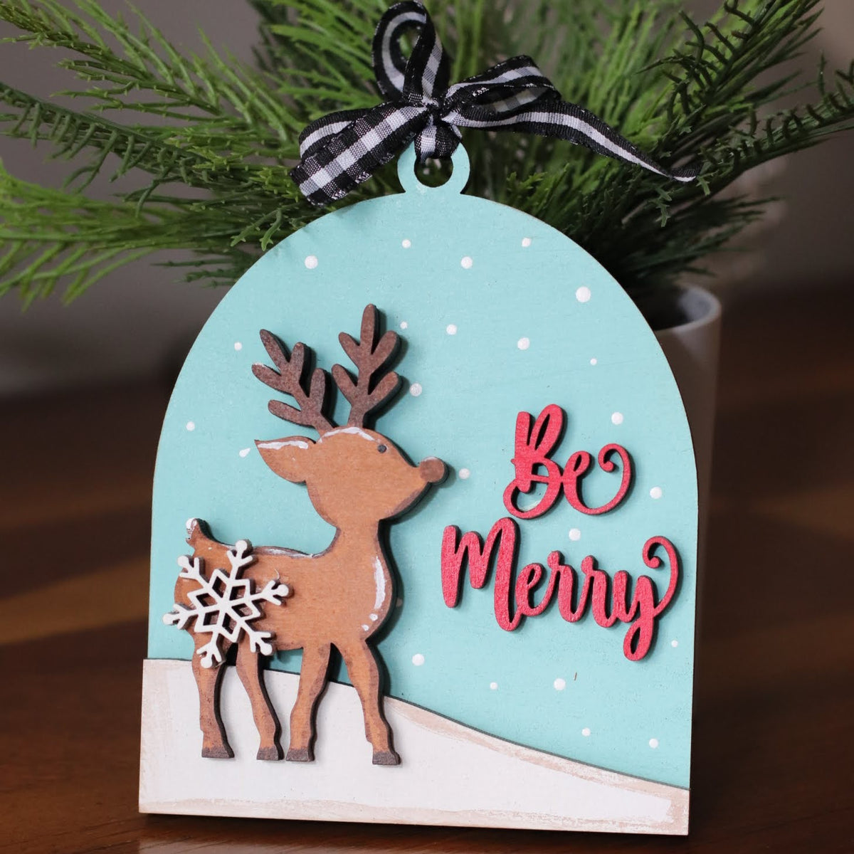 A handmade Christmas ornament with a die-cut arch shape, featuring a brown reindeer with antlers, a white snowflake, a light blue background with white dots, red 'Be Merry' script text, and a black-and-white gingham ribbon bow at the top. The ornament is displayed on a wooden surface with green pine branches in the background.