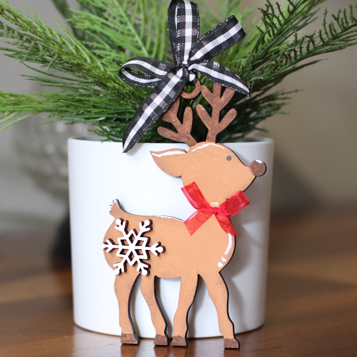 reindeer ornament with a black-and-white plaid ribbon bow, and a wooden reindeer cutout with a red bow and white snowflake detail, set on a wooden surface.
