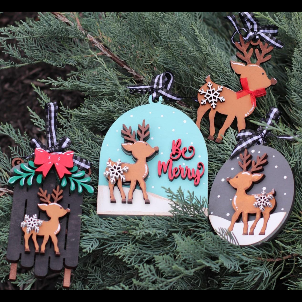 Three wooden Christmas ornaments with reindeer designs hanging on a green evergreen tree. One ornament is black with a red bow and green leaves, one is light blue with 'Be Merry' in red script, and one is gray with a white snow scene.