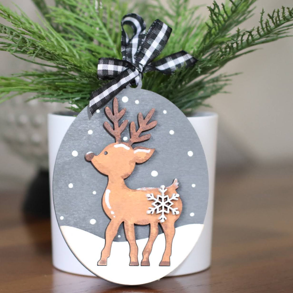 A decorative oval Christmas ornament featuring a die-cut reindeer with antlers and a white snowflake on its side, hanging from a black-and-white gingham ribbon bow. The ornament is displayed in front of a white candle holder with green pine branches on a wooden surface.